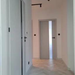 Korytarz z dwoma parami szarych drzwi wewnętrznych z czarnymi klamkami i oświetleniem szynowym, parkiet ułożony w jodełkę.