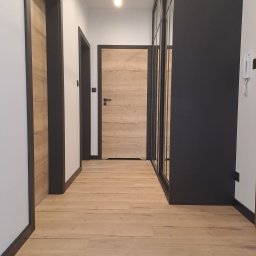 DAMAR PODŁOGI I DRZWI - Korytarz z drewnianą podłogą, dwiema parami drzwi z ciemnymi ościeżnicami i przesuwną szafą z czarnymi ramami.