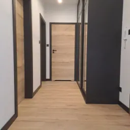 Korytarz z drewnianą podłogą, dwiema parami drzwi z ciemnymi ościeżnicami i przesuwną szafą z czarnymi ramami.