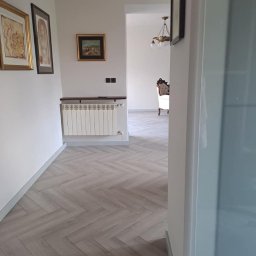 DAMAR PODŁOGI I DRZWI - Korytarz z podłogą ułożoną w jodełkę, widok zza uchylonych drzwi, w tle grzejnik pod trzema obrazami na ścianie i salon z ozdobnym żyrandolem i fotelem.