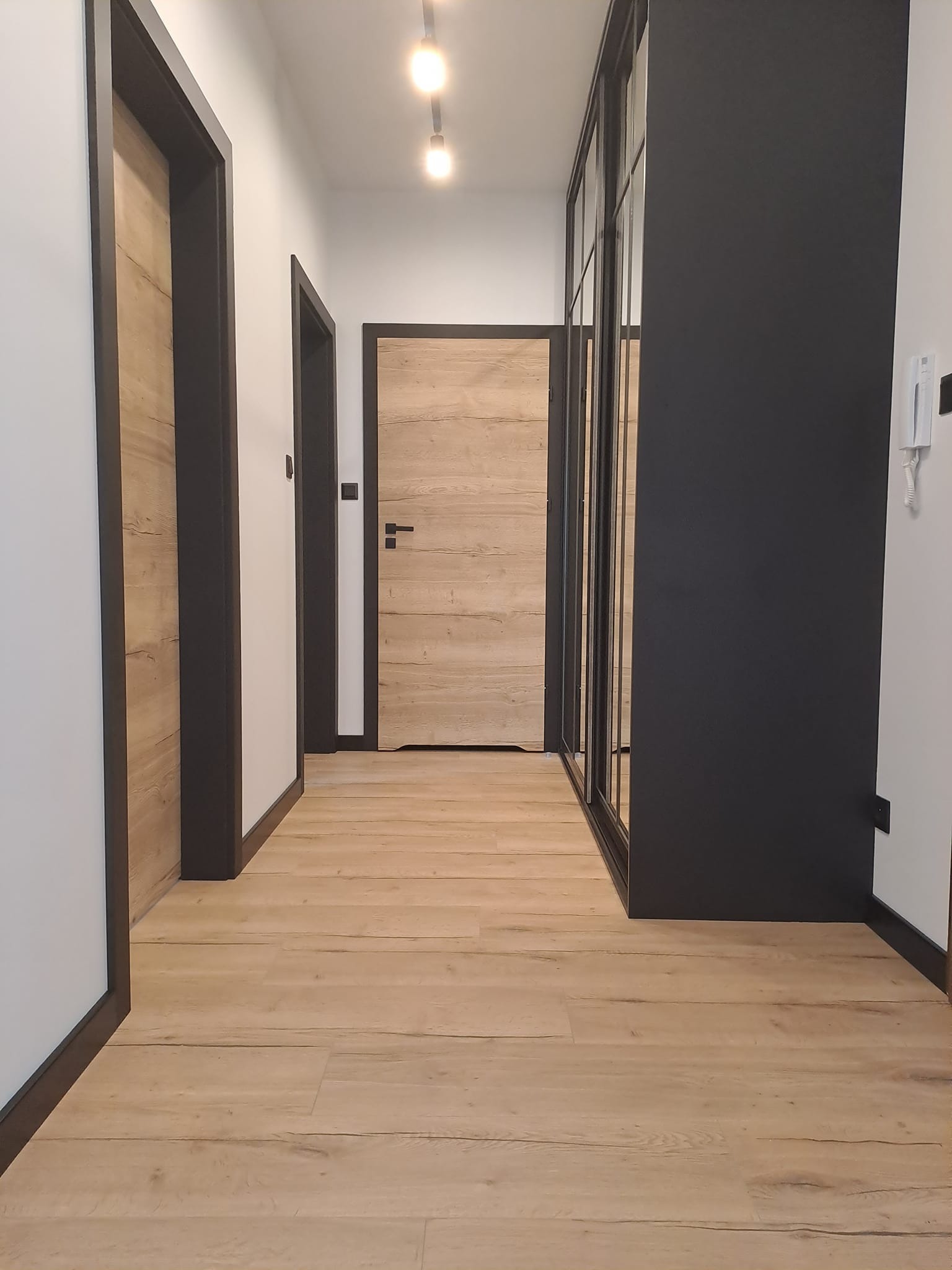 Korytarz z drewnianą podłogą, dwiema parami drzwi z ciemnymi ościeżnicami i przesuwną szafą z czarnymi ramami.