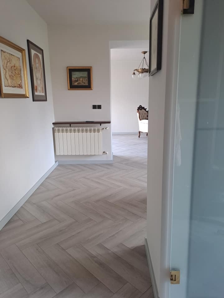 Korytarz z podłogą ułożoną w jodełkę, widok zza uchylonych drzwi, w tle grzejnik pod trzema obrazami na ścianie i salon z ozdobnym żyrandolem i fotelem.