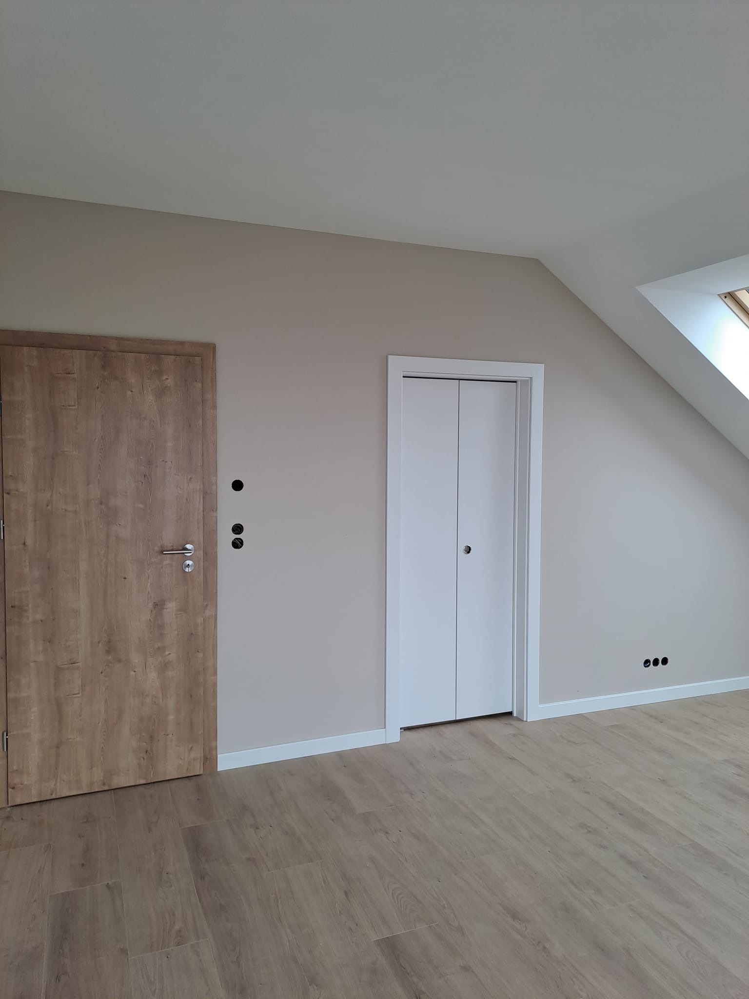 Pokój na poddaszu z drewnopodobną podłogą, brązowymi drzwiami i białymi drzwiami składanymi, widoczne gniazdka elektryczne i okno dachowe.