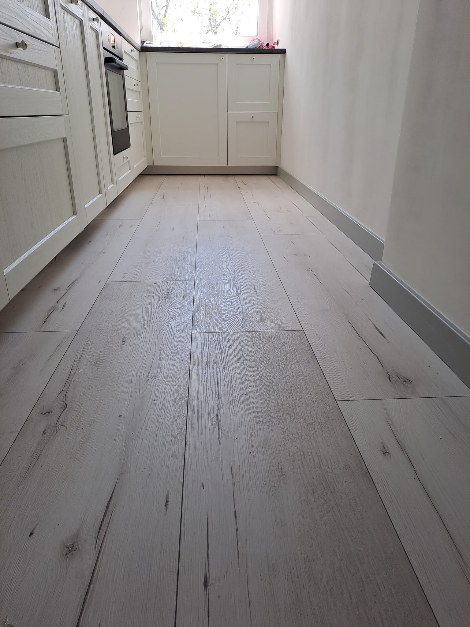 Ułożone panele podłogowe o jasnym odcieniu drewna w kuchni, widoczne białe szafki kuchenne i listwy przypodłogowe.