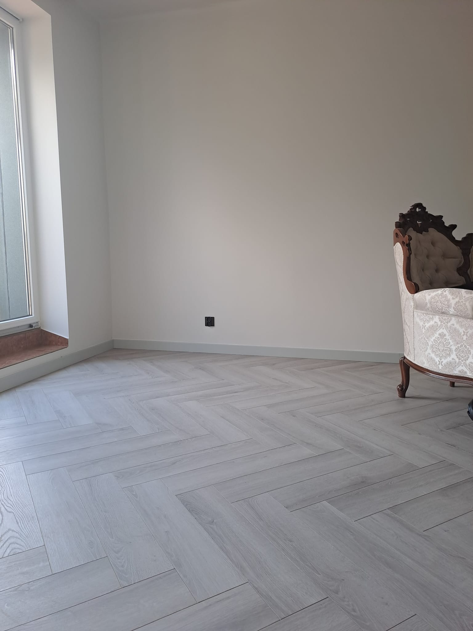 Jasnoszara podłoga ułożona we wzór jodełki francuskiej w pustym pokoju z oknem i częściowo widocznym, ozdobnym fotelem.
