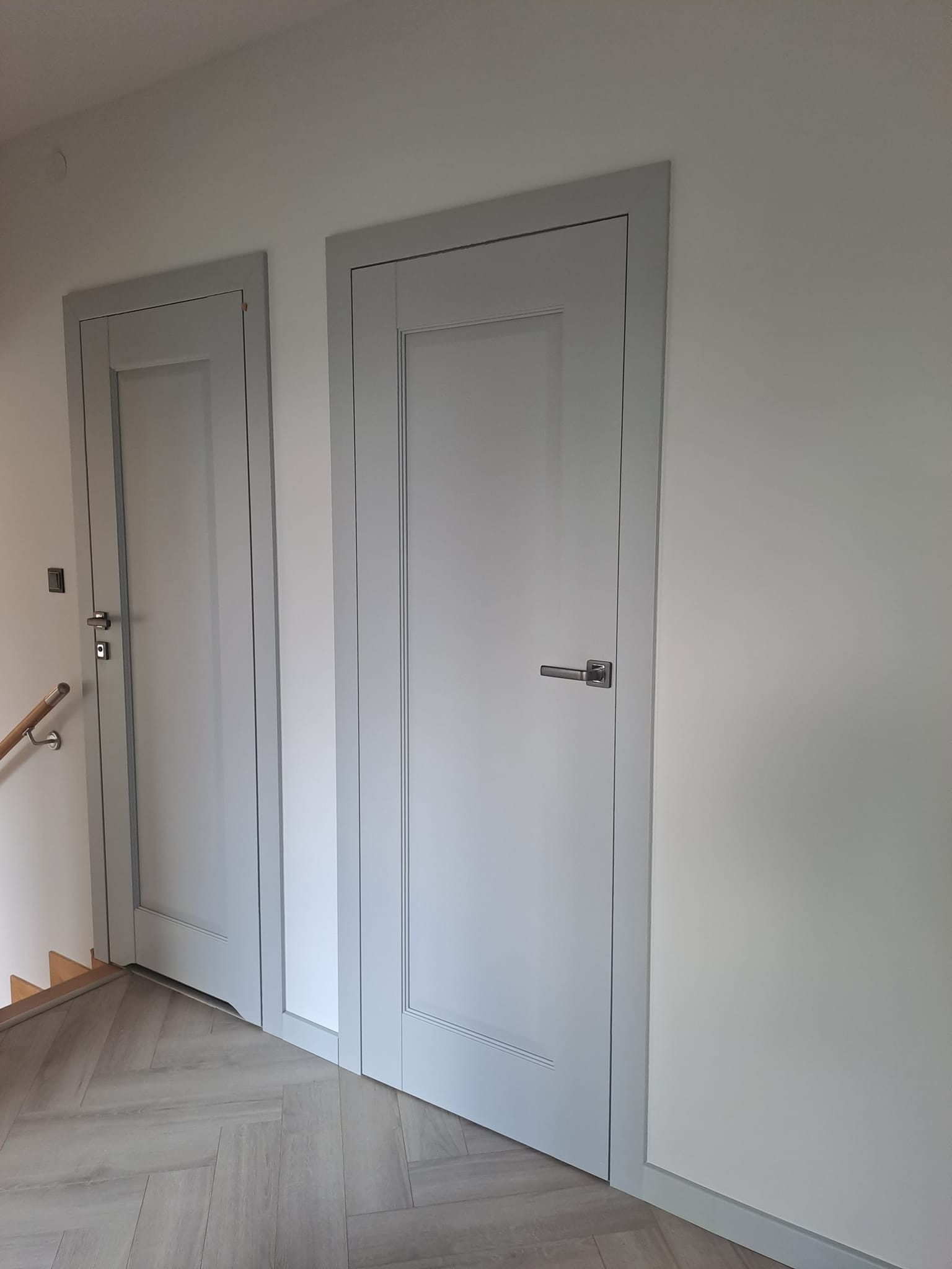 Dwa szare drzwi wewnętrzne z ramkami, zamontowane w korytarzu z podłogą w jodełkę, widoczne fragmenty schodów z drewnianą poręczą.