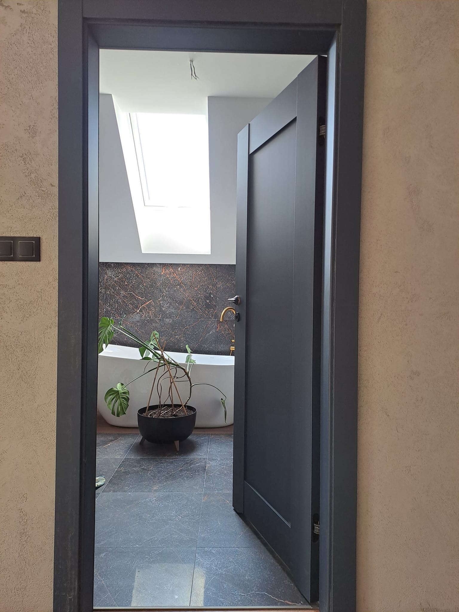 Ciemnoszare drzwi wewnętrzne uchylone, widok na łazienkę z oknem dachowym, wanną wolnostojącą i płytkami imitującymi marmur, doniczkowa monstera na podłodze.