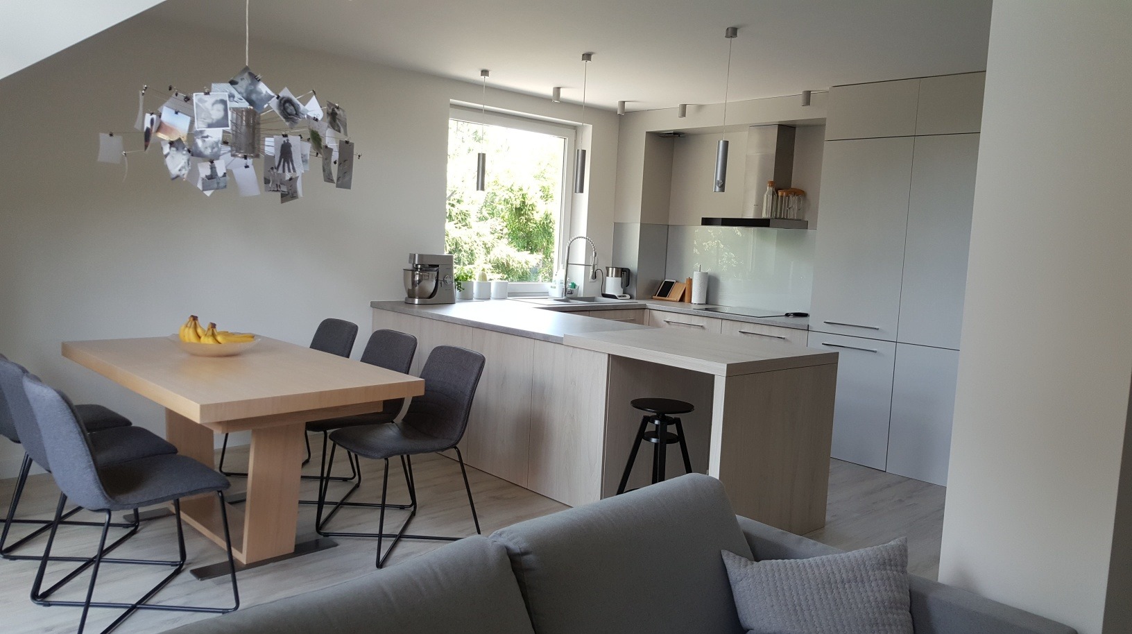 Jasna kuchnia w stylu skandynawskim z drewnianym stołem, szarymi krzesłami i lampą ozdobioną zdjęciami, na stole misa z bananami.