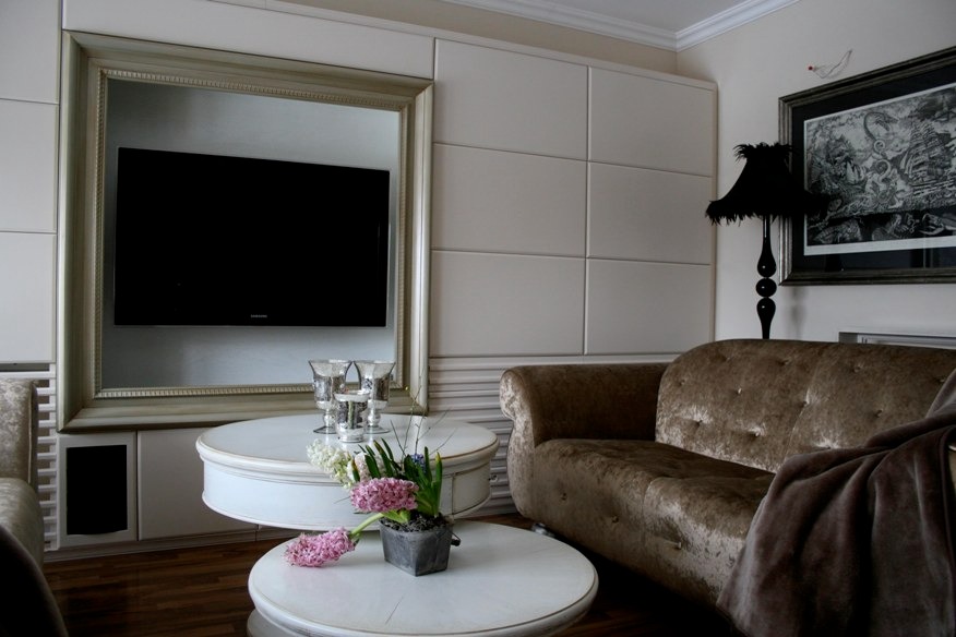 Salon w odcieniach beżu z telewizorem w lustrzanej ramie, okrągłymi stolikami z kwiatami i aksamitną sofą pod czarną lampą.