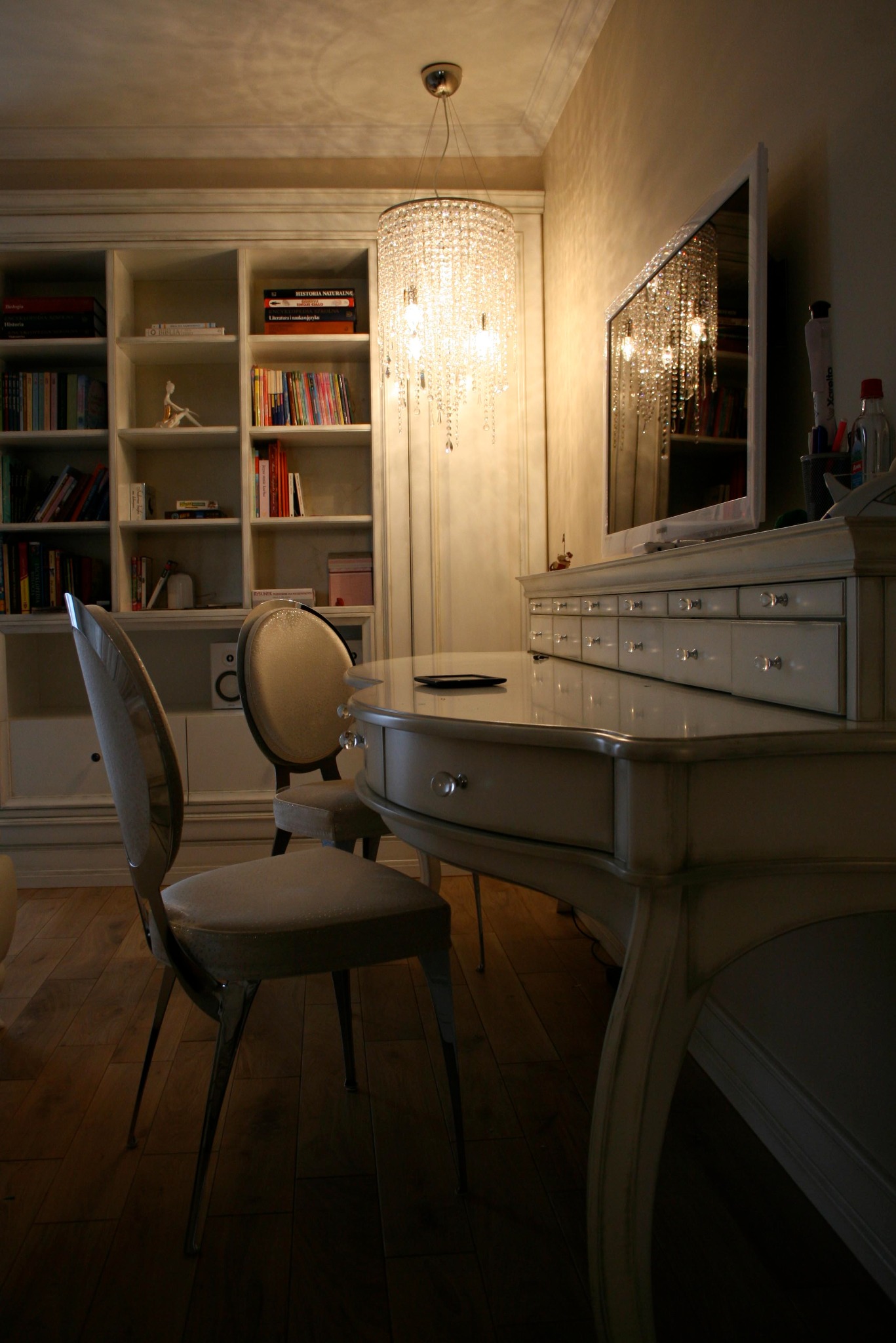 Eleganckie biurko z szufladami z kryształowymi gałkami, krzesła w stylu vintage, kryształowy żyrandol i regał z książkami w tle, oświetlone ciepłym światłem.