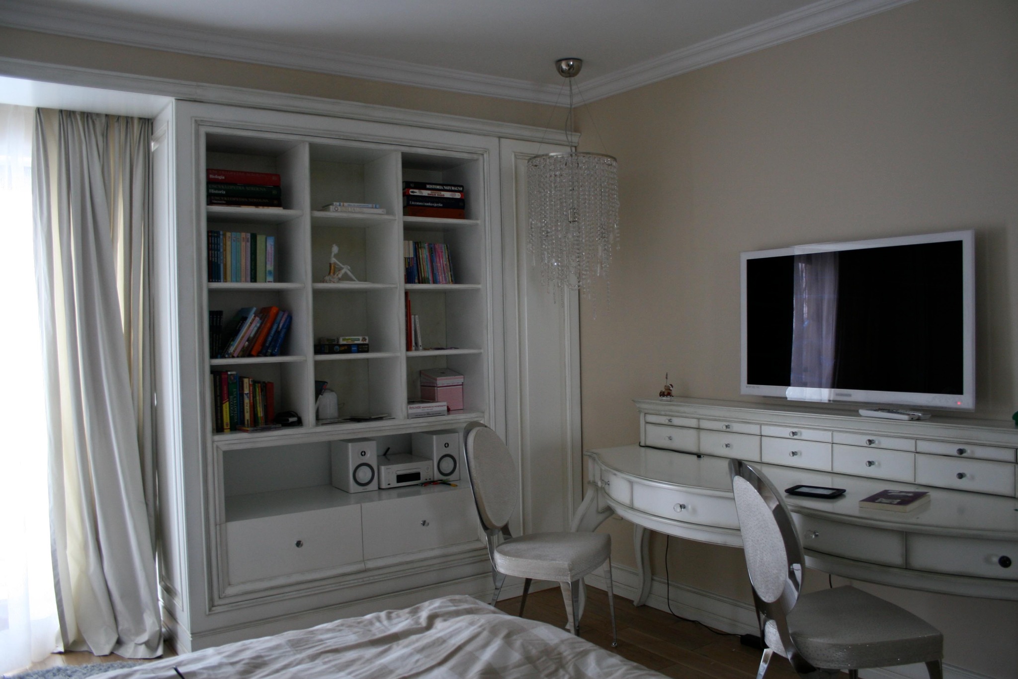 Jasna sypialnia z białymi meblami: regał z książkami, toaletka z telewizorem i kryształowy żyrandol. Dwa krzesła przy toaletce.