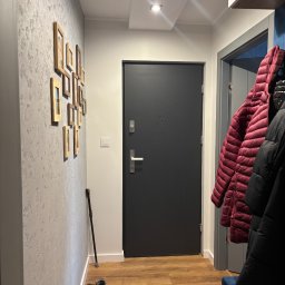 AD-BUD Adrian Wcisło - Wnętrze przedpokoju z szarą teksturowaną ścianą, na której wiszą ramki ze zdjęciami, ciemnoszare drzwi wejściowe, drewnopodobna podłoga, wieszak z kurtkami i siedzisko z niebieską tapicerką.