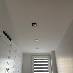 AD-BUD Adrian Wcisło - Biały sufit z trzema kwadratowymi, wpuszczanymi lampami LED w rzędzie, widok z dołu na tle białych szafek kuchennych i okna z żaluzjami.