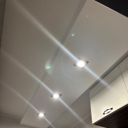 AD-BUD Adrian Wcisło - Biały, gładki sufit z wpuszczonymi, kwadratowymi lampami LED, widoczny fragment białej szafki kuchennej z czarnym uchwytem, oświetlenie punktowe.