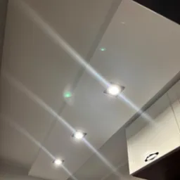 Biały, gładki sufit z wpuszczonymi, kwadratowymi lampami LED, widoczny fragment białej szafki kuchennej z czarnym uchwytem, oświetlenie punktowe.