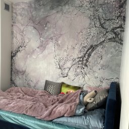 AD-BUD Adrian Wcisło - Sypialnia z łóżkiem i dekoracyjną tapetą w odcieniach szarości i różu z motywem kwitnących gałęzi, uzupełniona o tekstylia w pastelowych kolorach.