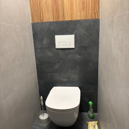 AD-BUD Adrian Wcisło - Nowoczesna toaleta w szaro-czarnej kolorystyce z drewnianą szafką nad spłuczką, widoczne akcesoria łazienkowe: szczotka do WC, płyn do czyszczenia i papier toaletowy.