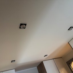 AD-BUD Adrian Wcisło - Biały sufit z zamontowanymi czterema czarnymi, kwadratowymi oprawami oświetleniowymi typu spot, widoczne fragmenty zabudowy meblowej.