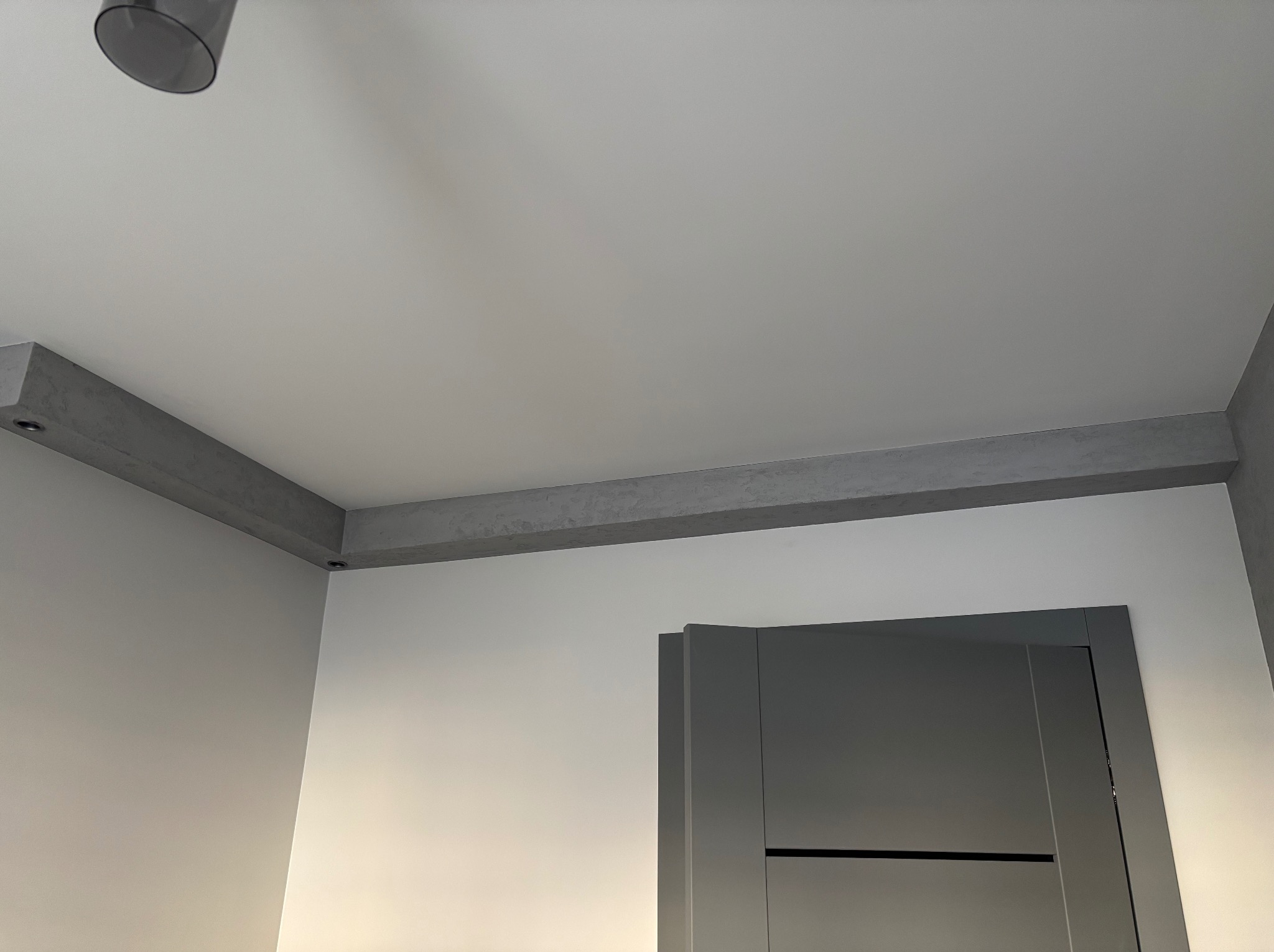 Nowoczesne wnętrze z minimalistycznym sufitem, szarymi belkami dekoracyjnymi z wbudowanym oświetleniem punktowym i częściowo widocznymi szarymi drzwiami.
