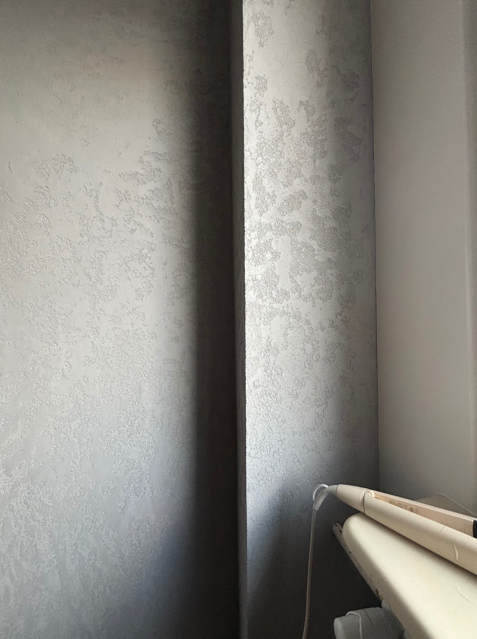 Ściana z dekoracyjną, szarą strukturą tynku obok gładkiej ściany i fragmentu deski do prasowania.