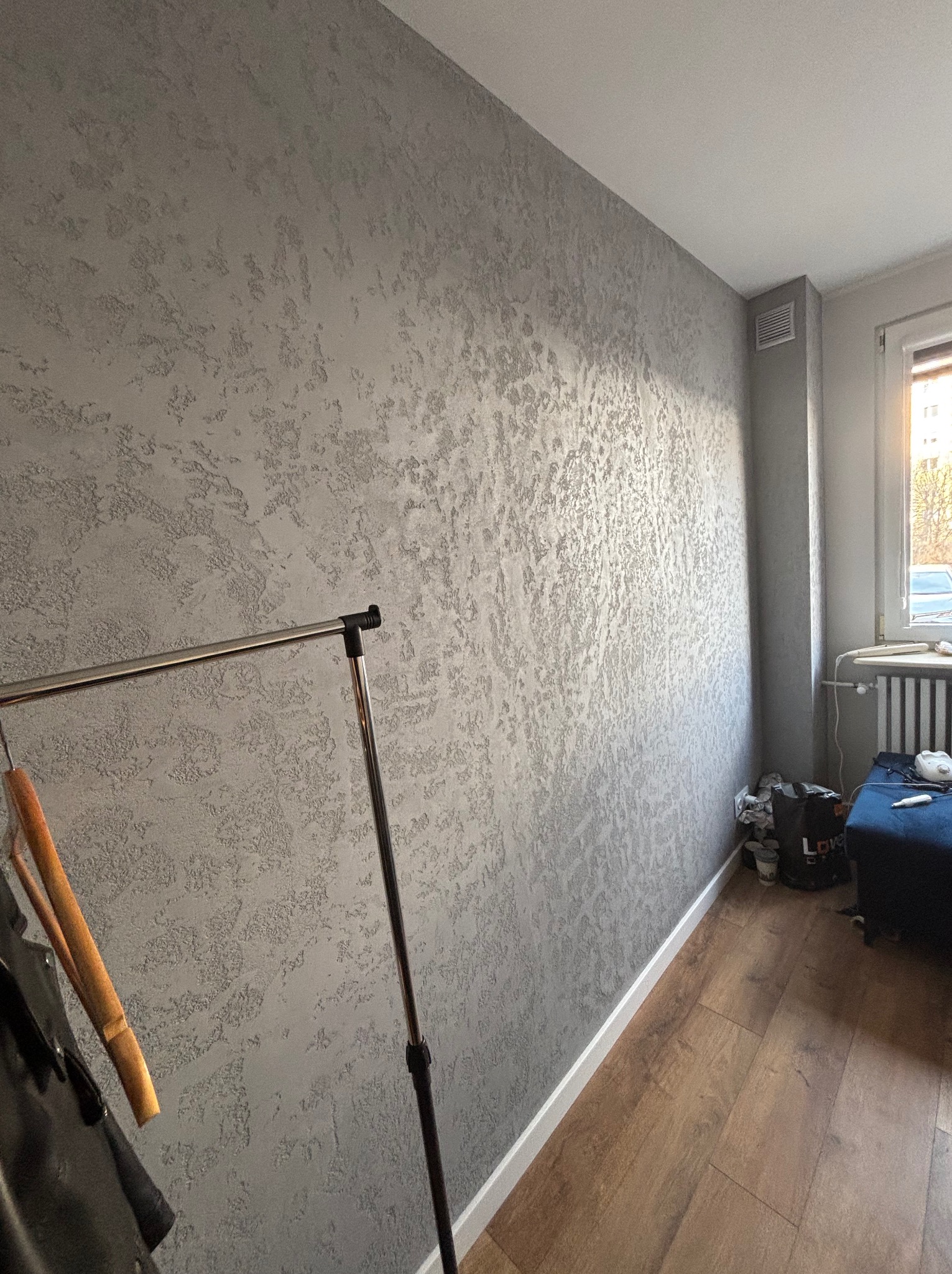 Ściana w pokoju pokryta dekoracyjnym tynkiem w odcieniach szarości, z widoczną fakturą i metalicznym połyskiem, obok drewniana podłoga i fragment wieszaka na ubrania.