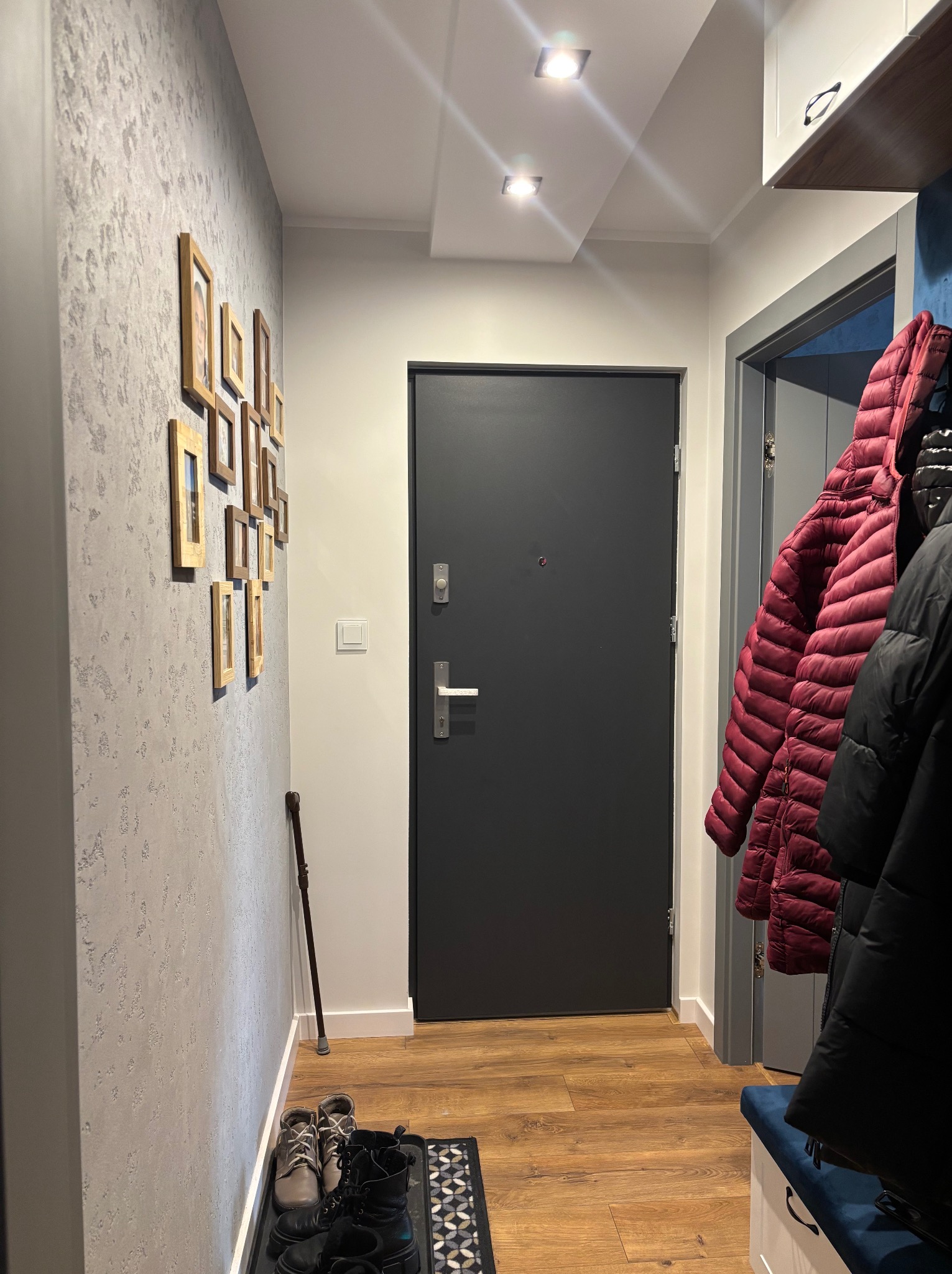 Wnętrze przedpokoju z szarą teksturowaną ścianą, na której wiszą ramki ze zdjęciami, ciemnoszare drzwi wejściowe, drewnopodobna podłoga, wieszak z kurtkami i siedzisko z niebieską tapicerką.