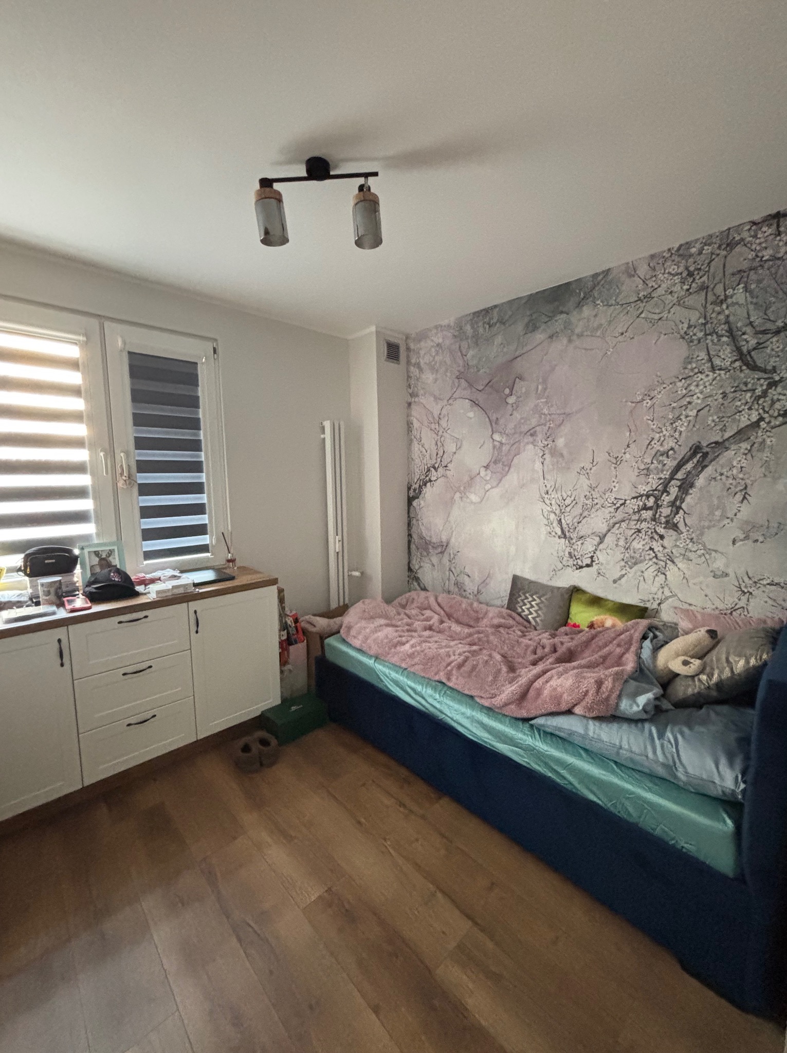 Pokój po odświeżeniu: tapeta w kwiaty na ścianie za łóżkiem z turkusową narzutą i różowym kocem, białe szafki z ciemnymi uchwytami wzdłuż ściany, drewniana podłoga.