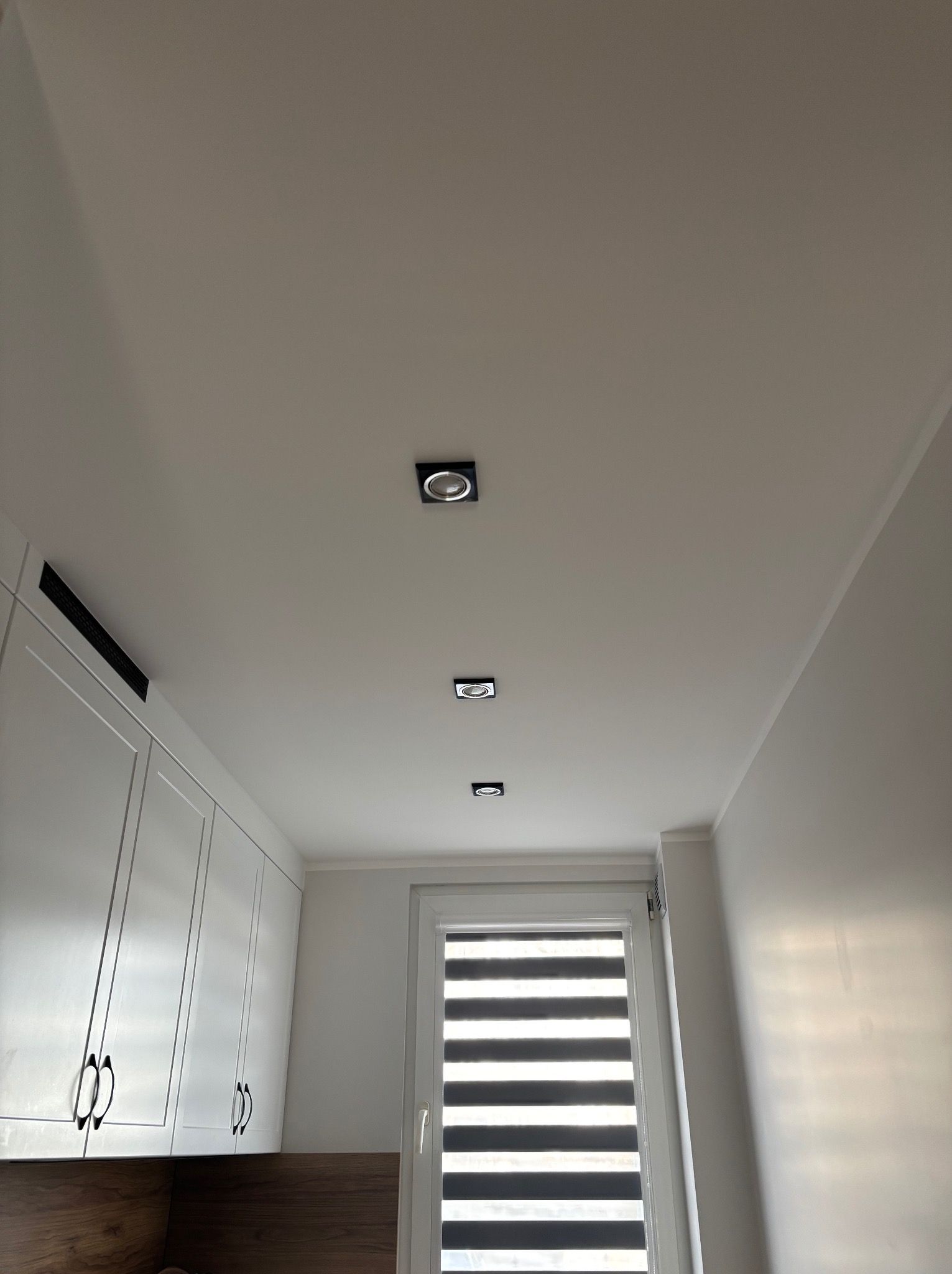 Biały sufit z trzema kwadratowymi, wpuszczanymi lampami LED w rzędzie, widok z dołu na tle białych szafek kuchennych i okna z żaluzjami.