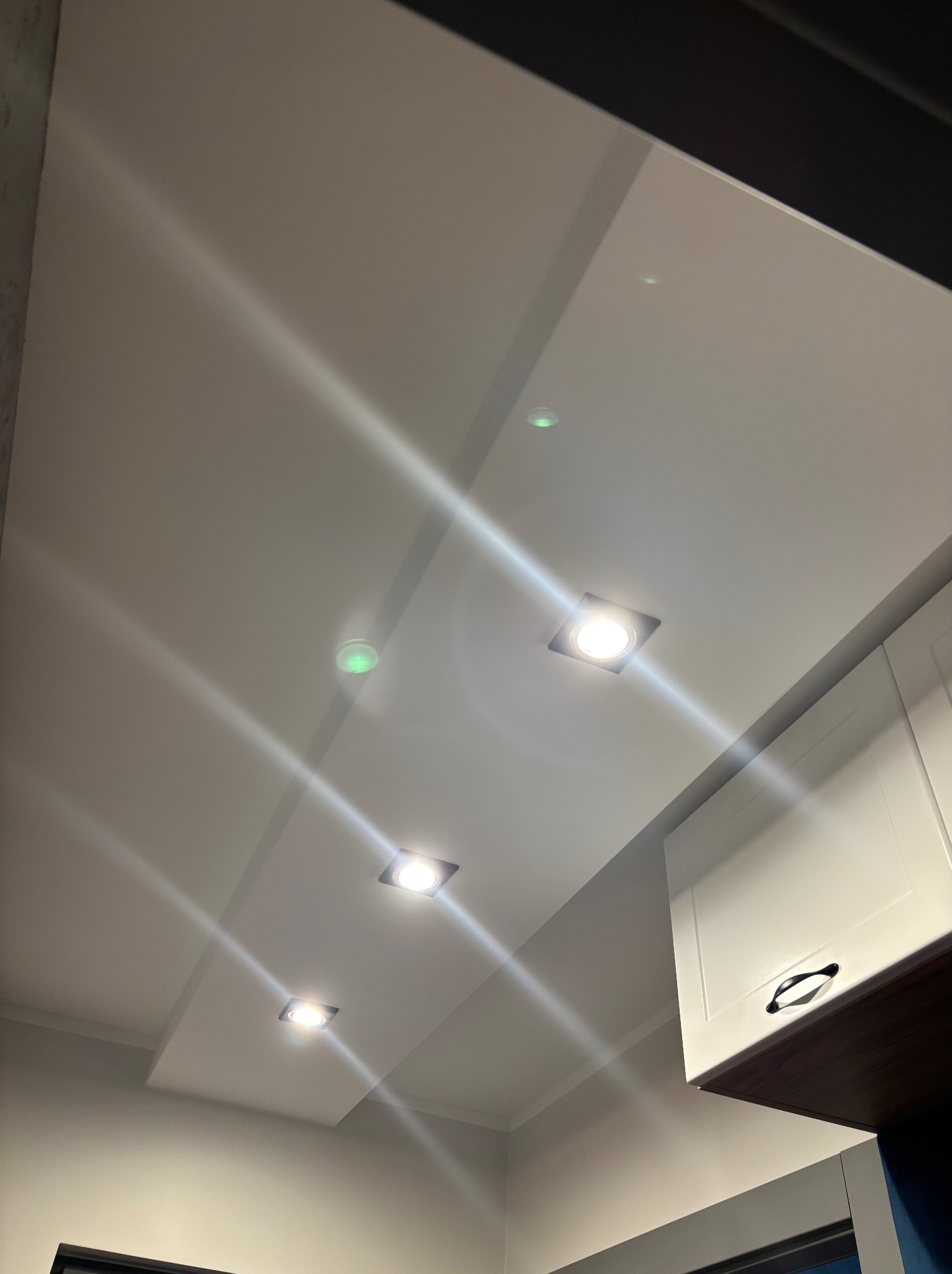 Biały, gładki sufit z wpuszczonymi, kwadratowymi lampami LED, widoczny fragment białej szafki kuchennej z czarnym uchwytem, oświetlenie punktowe.