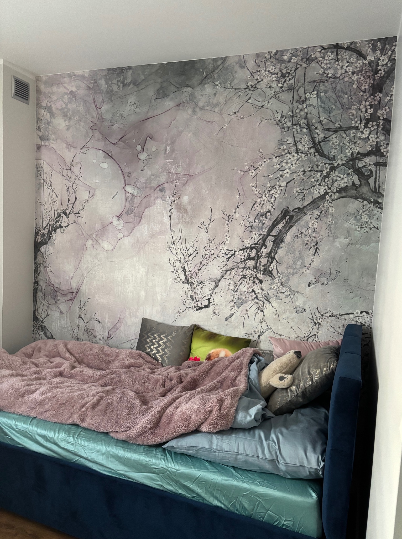Sypialnia z łóżkiem i dekoracyjną tapetą w odcieniach szarości i różu z motywem kwitnących gałęzi, uzupełniona o tekstylia w pastelowych kolorach.