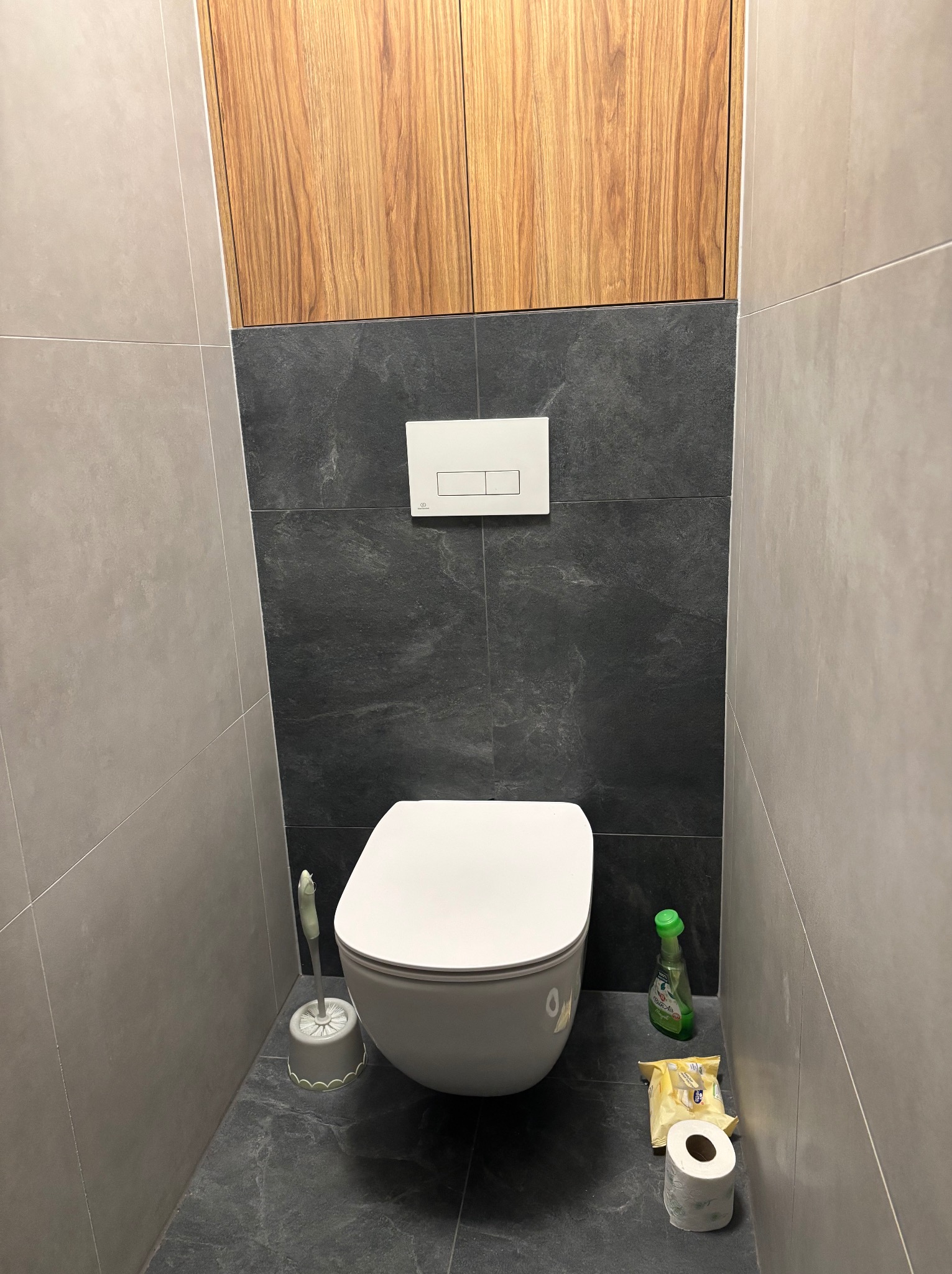 Nowoczesna toaleta w szaro-czarnej kolorystyce z drewnianą szafką nad spłuczką, widoczne akcesoria łazienkowe: szczotka do WC, płyn do czyszczenia i papier toaletowy.