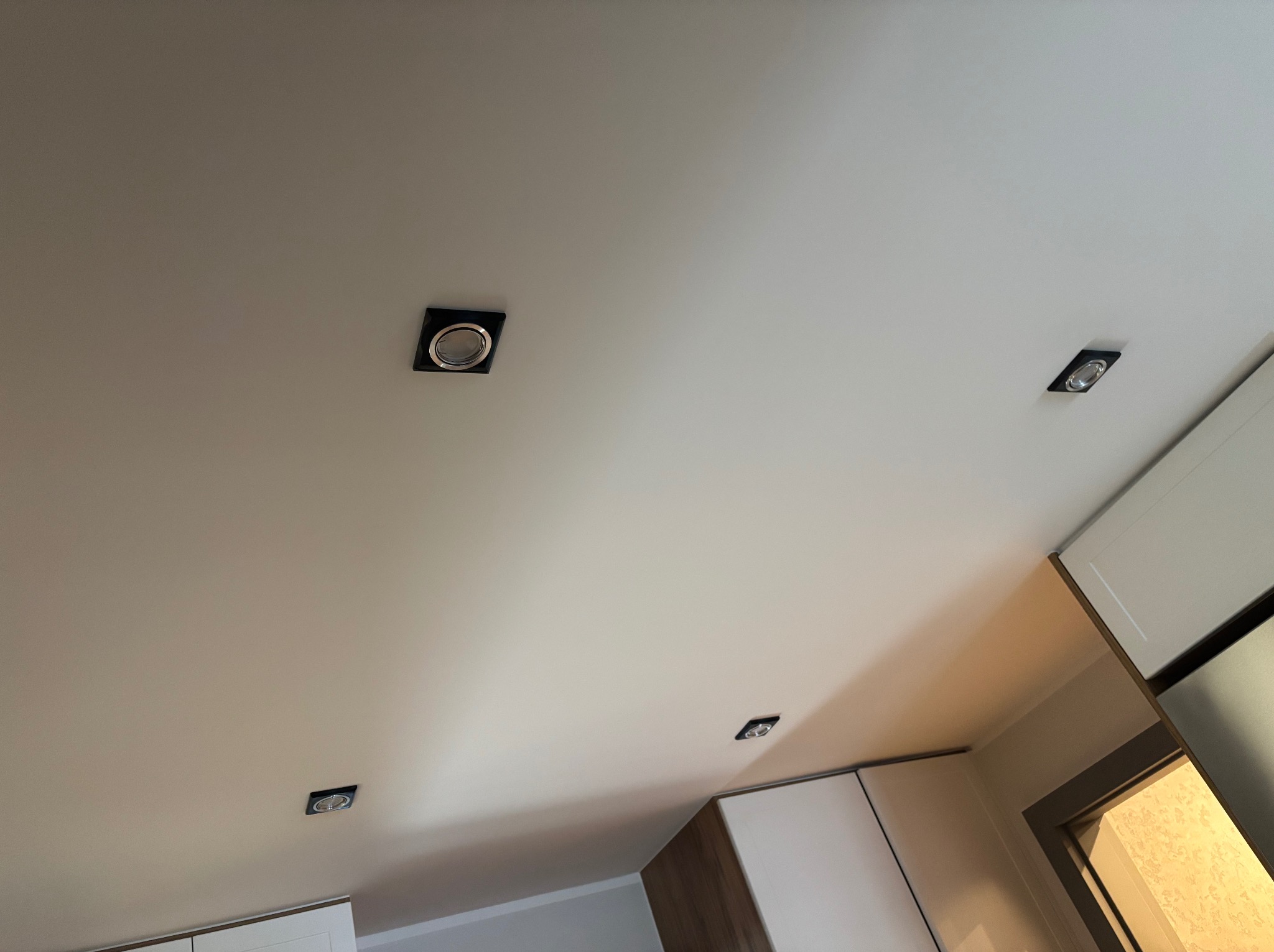 Biały sufit z zamontowanymi czterema czarnymi, kwadratowymi oprawami oświetleniowymi typu spot, widoczne fragmenty zabudowy meblowej.