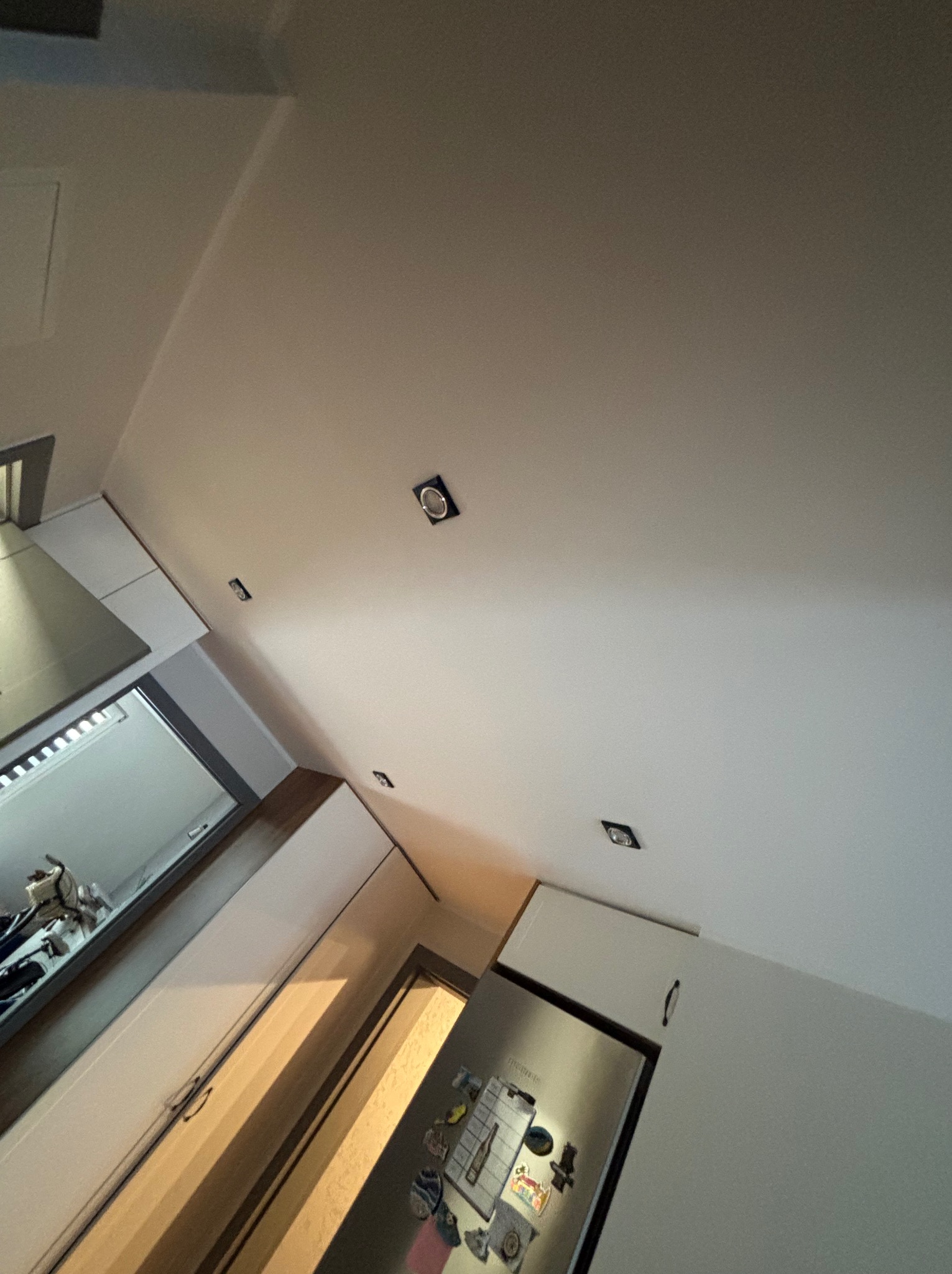 Nowoczesna kuchnia z białymi szafkami, srebrną lodówką, i oświetleniem punktowym w suficie, widziana pod nietypowym kątem.