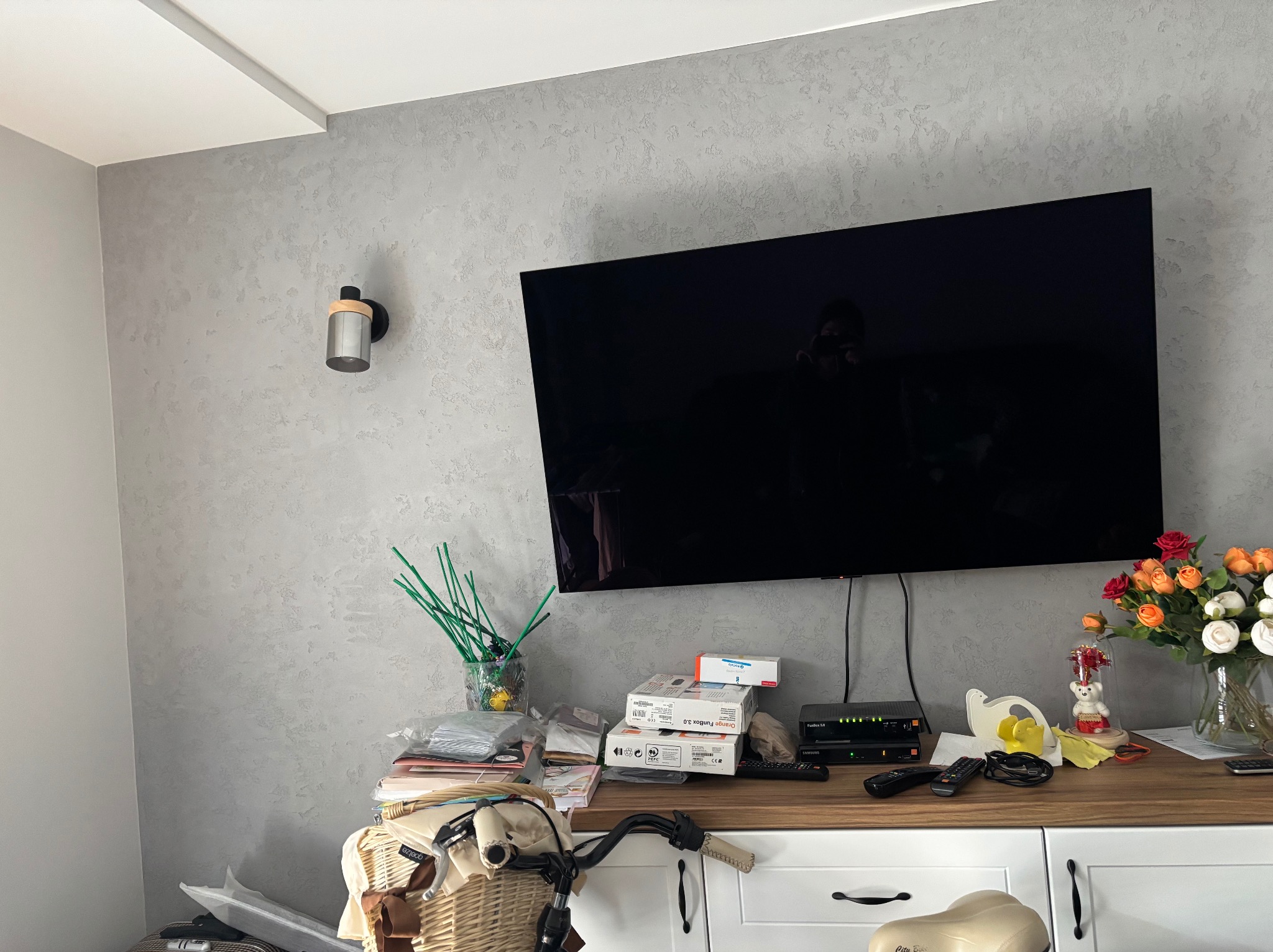 Ściana pokryta dekoracyjnym tynkiem w odcieniach szarości, z zamontowanym telewizorem i lampą ścienną, widoczny fragment białych mebli z rowerem i kwiatami na blacie.