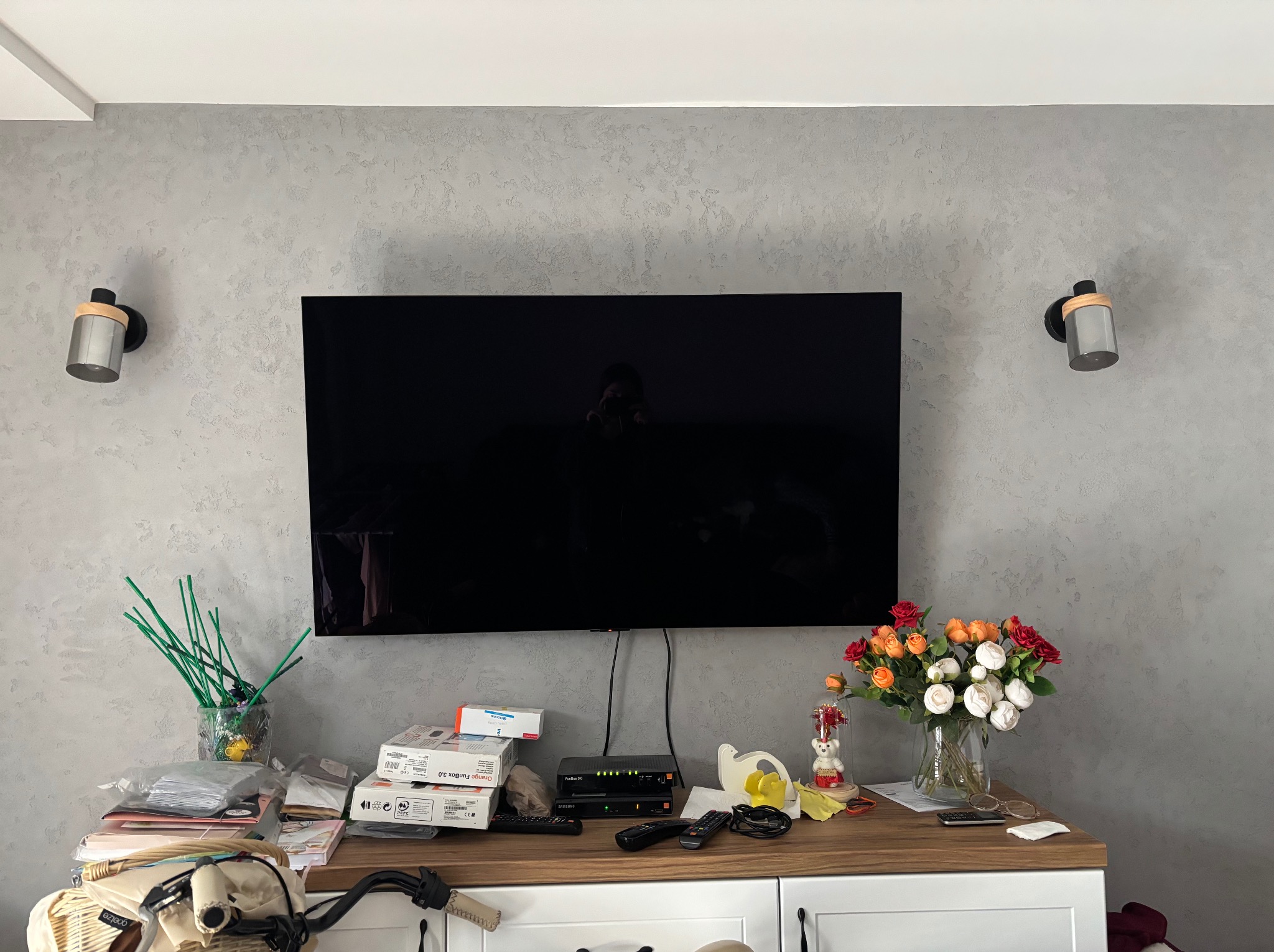 Ściana z dekoracyjnym tynkiem w odcieniach szarości, oświetlona przez dwa kinkiety z drewnianymi akcentami, nad komodą z telewizorem i dekoracjami.