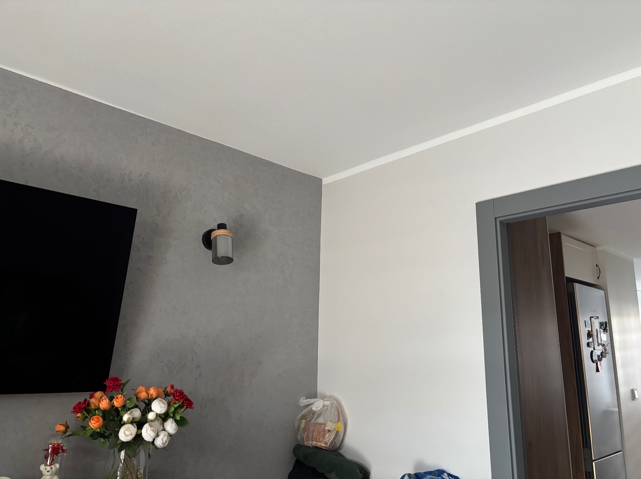 Szara ściana z dekoracyjnym tynkiem w salonie, biały sufit z listwą, widoczny fragment telewizora, bukiet kwiatów w wazonie i lampa ścienna.
