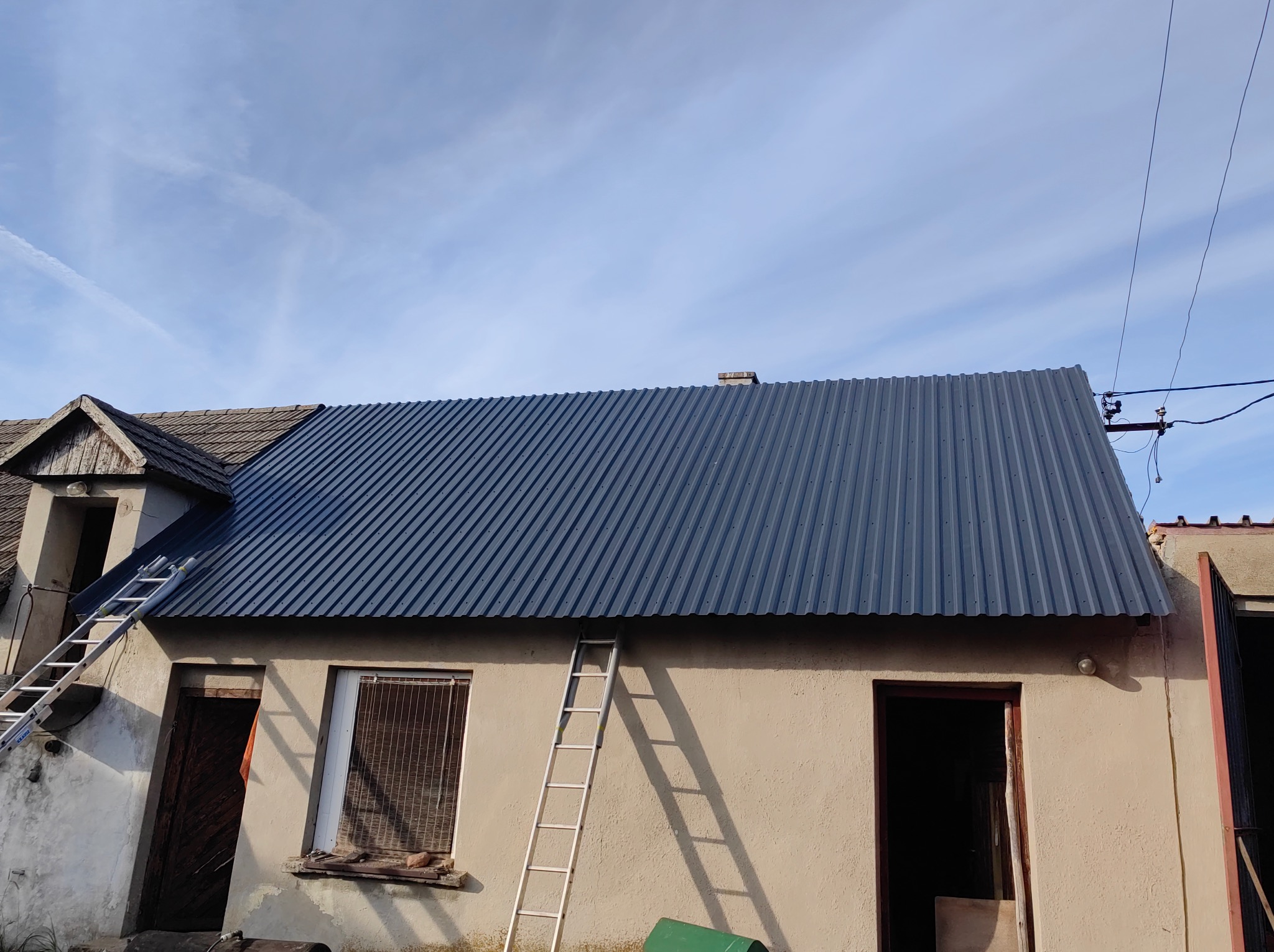Nowe, błyszczące, grafitowe pokrycie dachowe z blachy trapezowej na budynku gospodarczym, z drabiny opartymi o ścianę i dach, widoczne elementy starego dachu z dachówki.