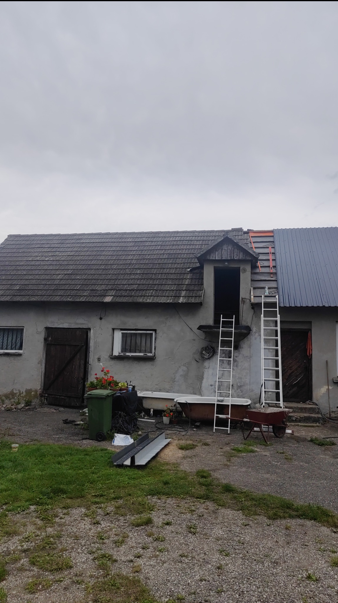 Dwukondygnacyjny budynek z szarą elewacją i częściowo wymienionym pokryciem dachu, widoczne dwie drabiny opierające się o budynek, przy budynku stoi wanna i taczka.