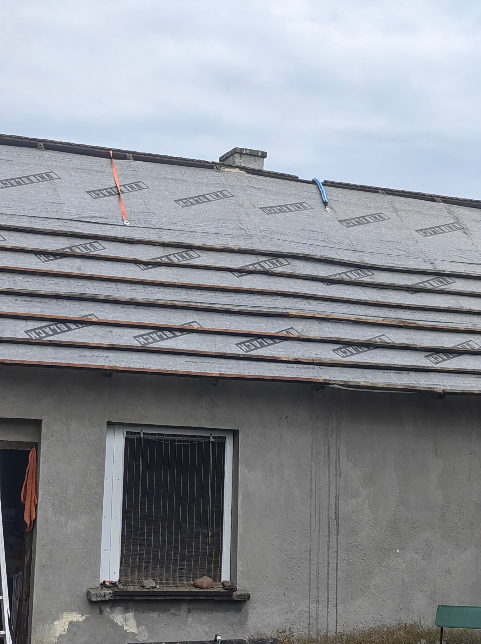 Szare pokrycie dachowe z widocznym logo UNI BLACK, zabezpieczone pomarańczowym i niebieskim pasem transportowym, na tle szarej elewacji budynku z oknem z kratą i otwartymi drzwiami.