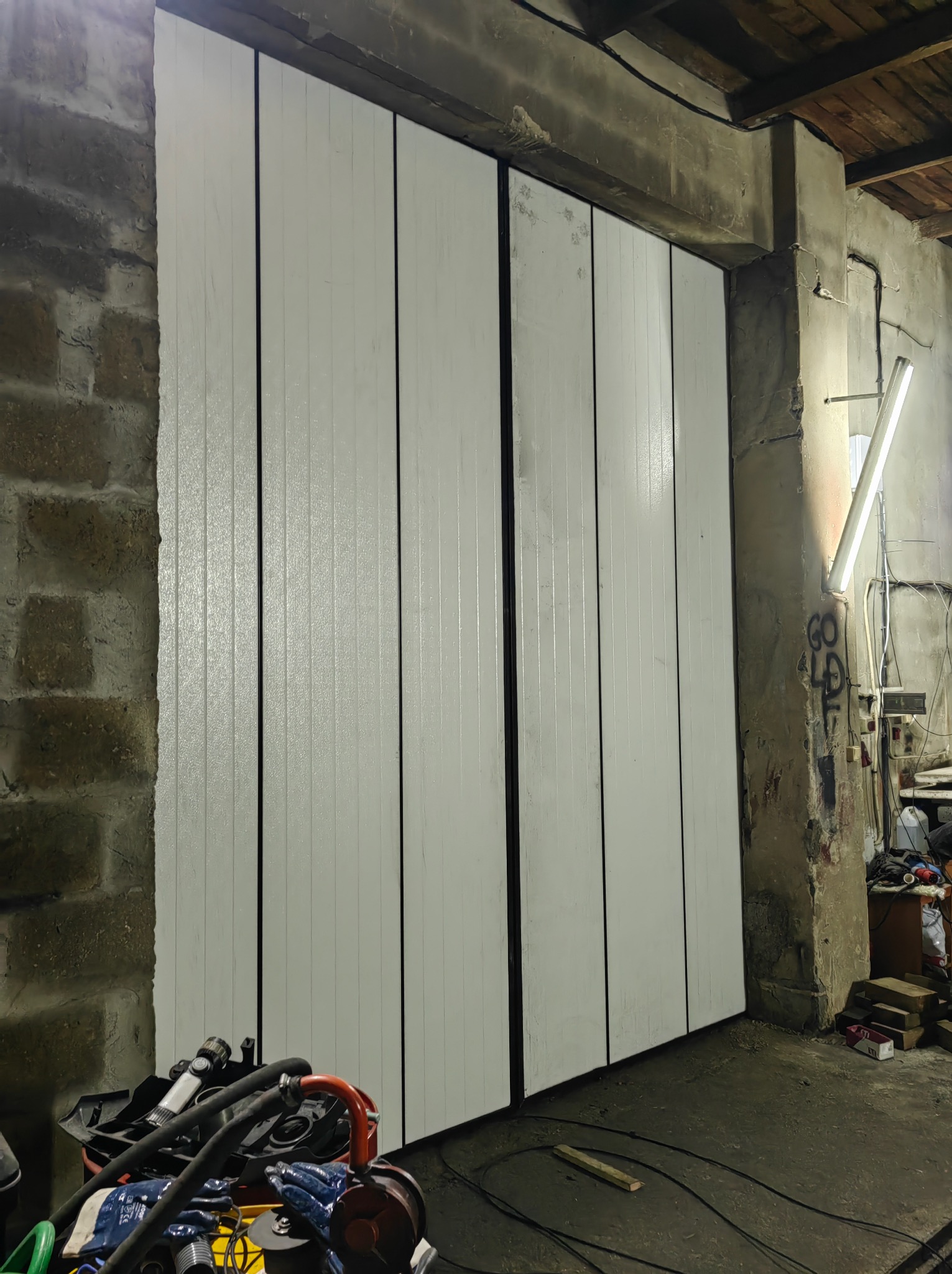 Białe, dwuskrzydłowe drzwi garażowe z pionowych desek, widoczne na tle surowej, ceglanej ściany i betonowego filaru z instalacją elektryczną.