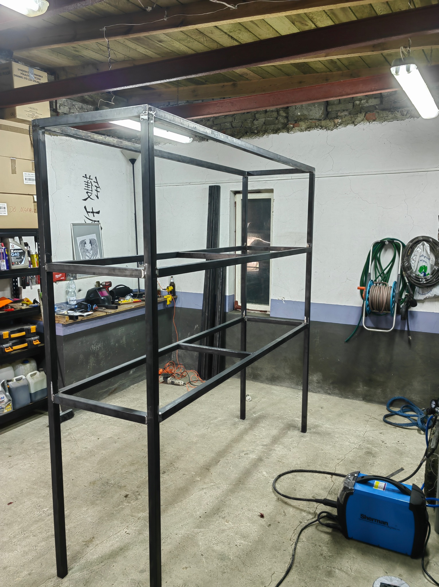 Spawana metalowa konstrukcja regału z profili zamkniętych, widoczna spawarka Sherman, warsztatowe otoczenie z narzędziami i akcesoriami.