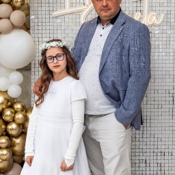 Artur Gębura - Portret dziewczynki w białej sukience komunijnej z tatą na tle ściany z dekoracją balonową i neonowym napisem 'Komunia Święta'.