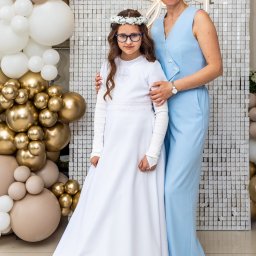 Artur Gębura - Dziewczynka w białej sukience komunijnej z mamą na tle dekoracji z balonów i błyszczącej ścianki, ujęcie w pełnej sylwetce, delikatne oświetlenie.