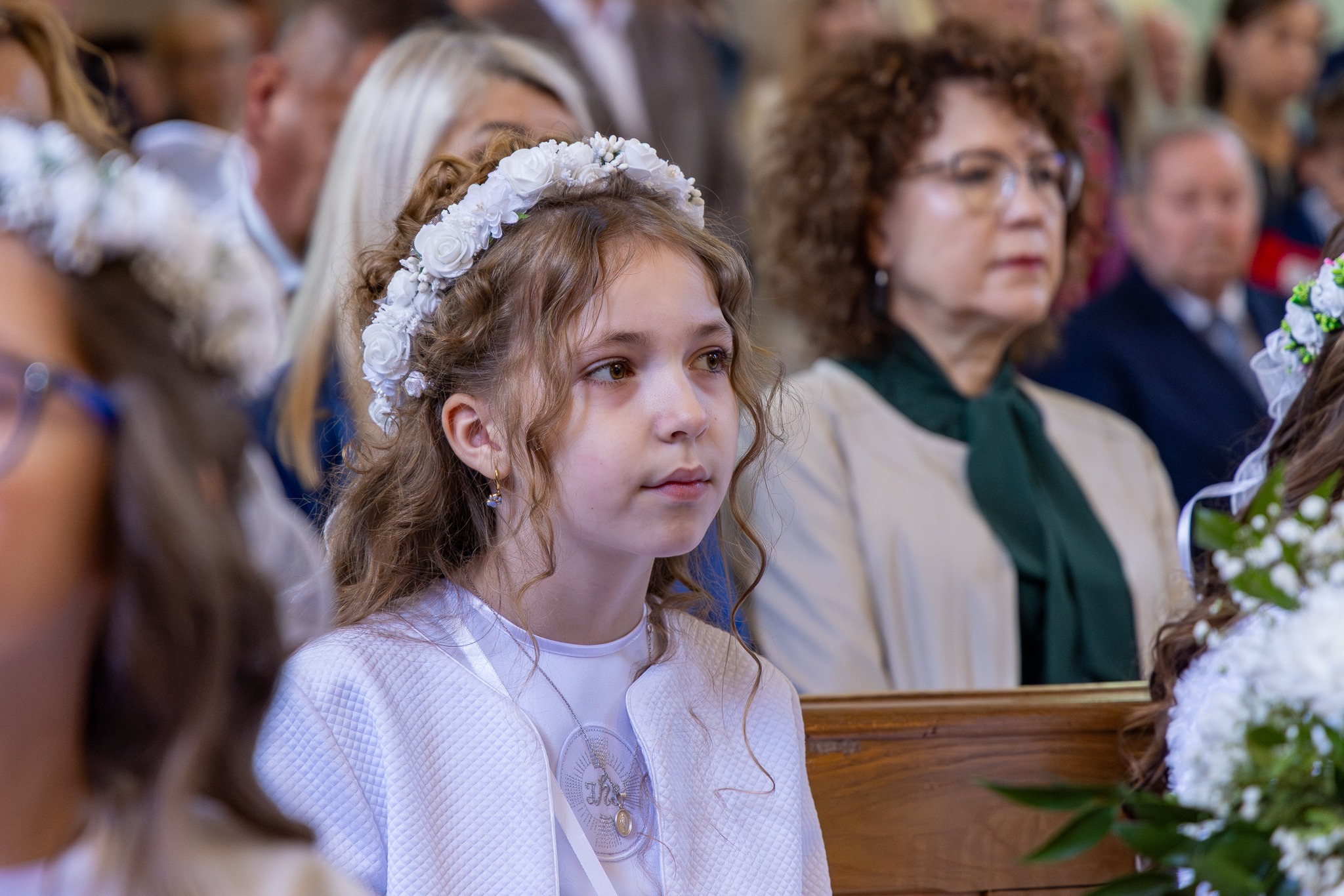 Dziewczynka w stroju komunijnym z wiankiem na głowie, siedząca w kościele podczas uroczystości, z delikatnym oświetleniem i widocznymi detalami stroju i fryzury.