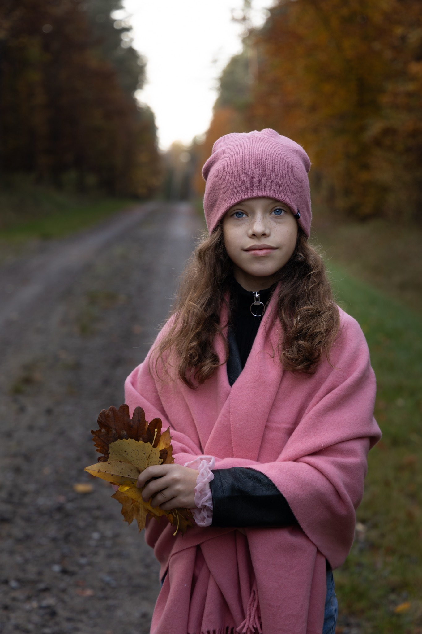Dziewczynka w różowej czapce i szalu pozuje z liśćmi na leśnej drodze. Jesienna sceneria, naturalne światło, portret w plenerze. Długie, kręcone włosy.