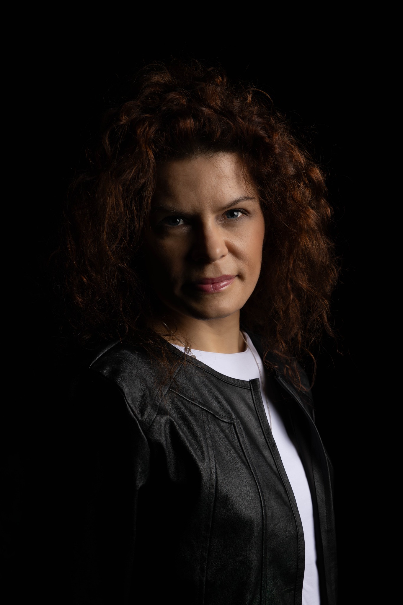 Portret kobiety o rudych kręconych włosach, ubrana w czarną skórzaną kurtkę na białej koszulce, na ciemnym tle. Subtelne oświetlenie podkreśla rysy twarzy.