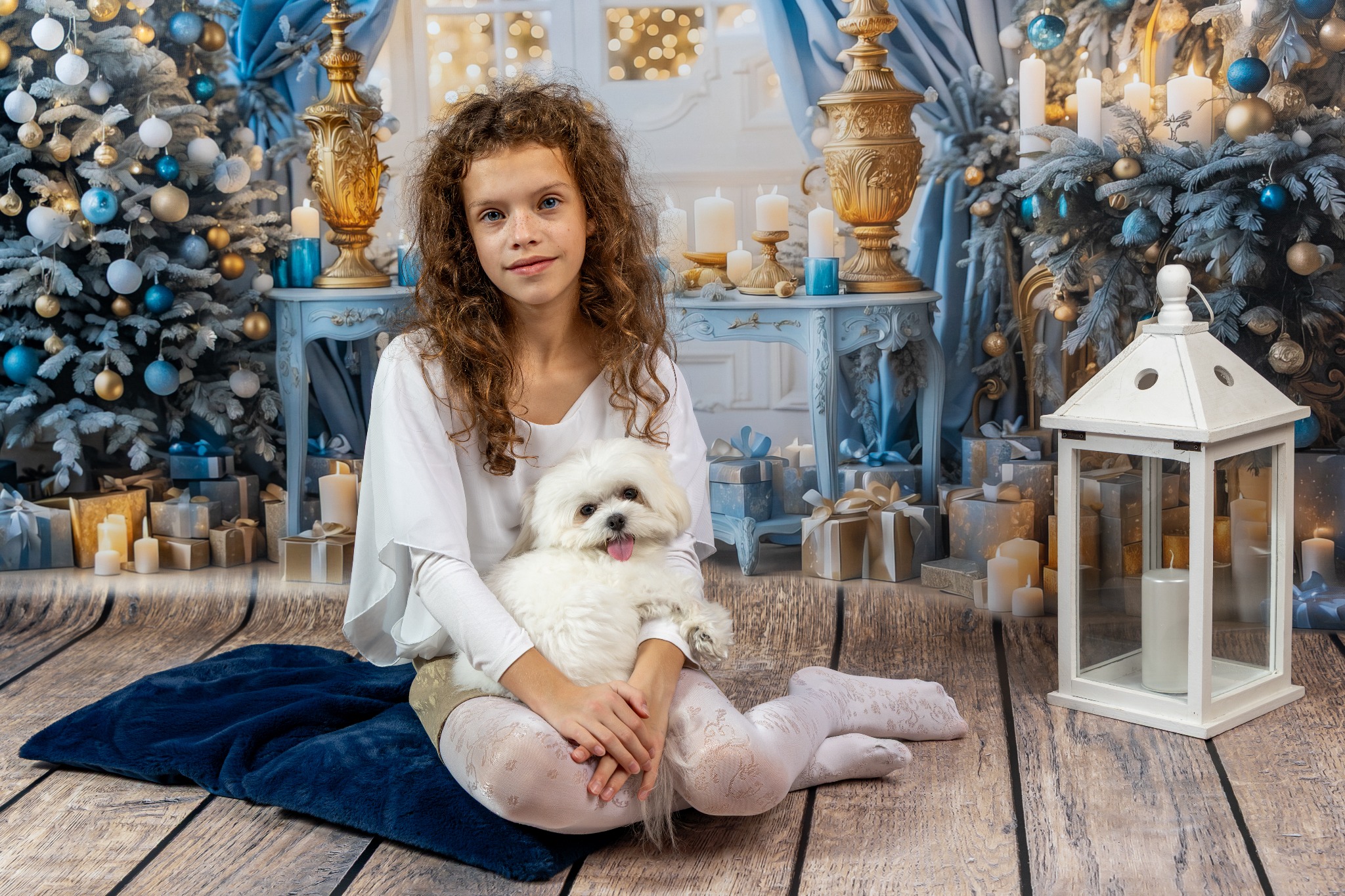 Dziewczynka z kręconymi włosami trzyma białego pieska na tle świątecznej scenografii z choinkami, prezentami i świecami w ciepłej tonacji.