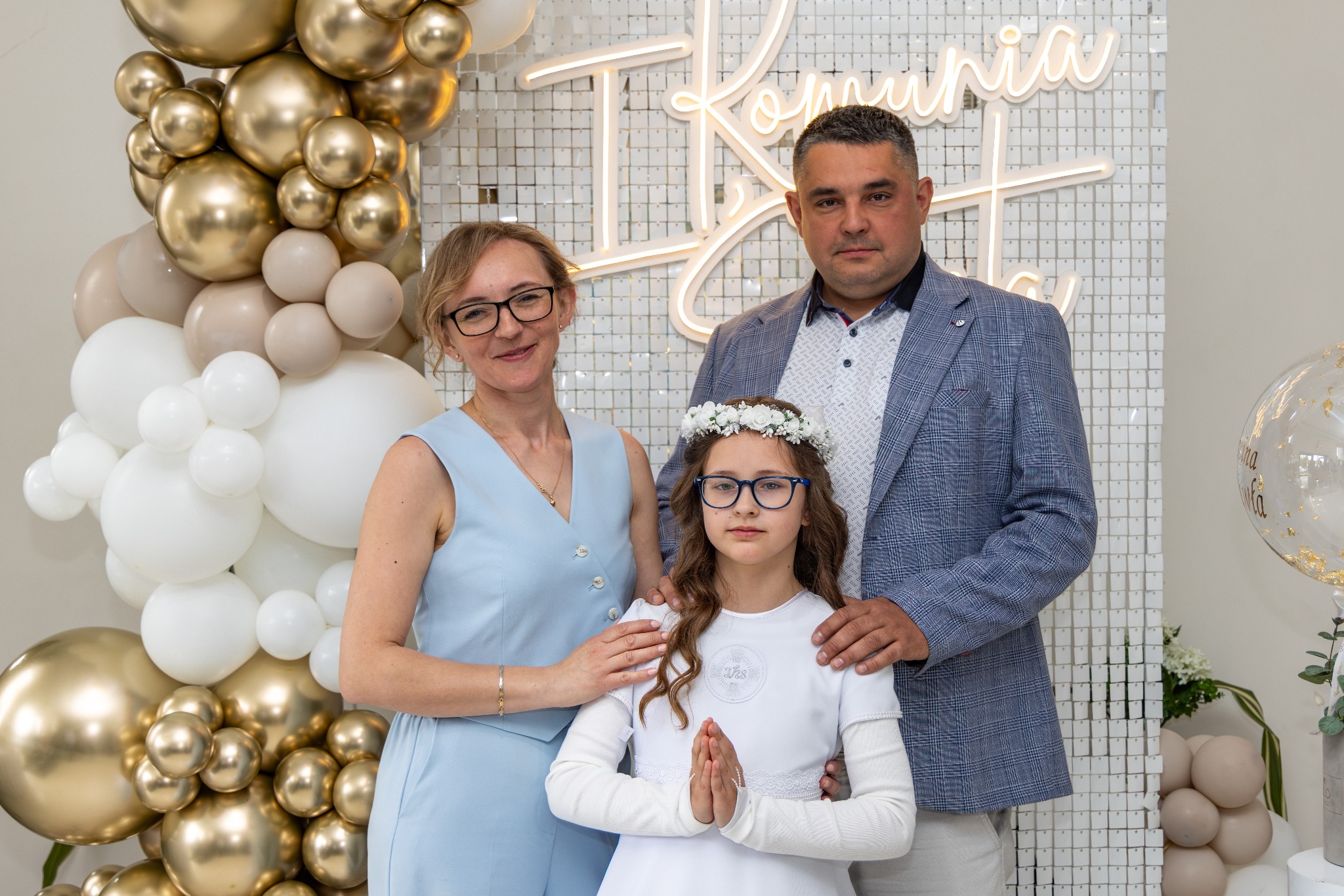 Rodzinne zdjęcie z Pierwszej Komunii Świętej: dziewczynka w albie, rodzice obok, dekoracje z balonów i błyszcząca ścianka w tle, ciepłe oświetlenie.