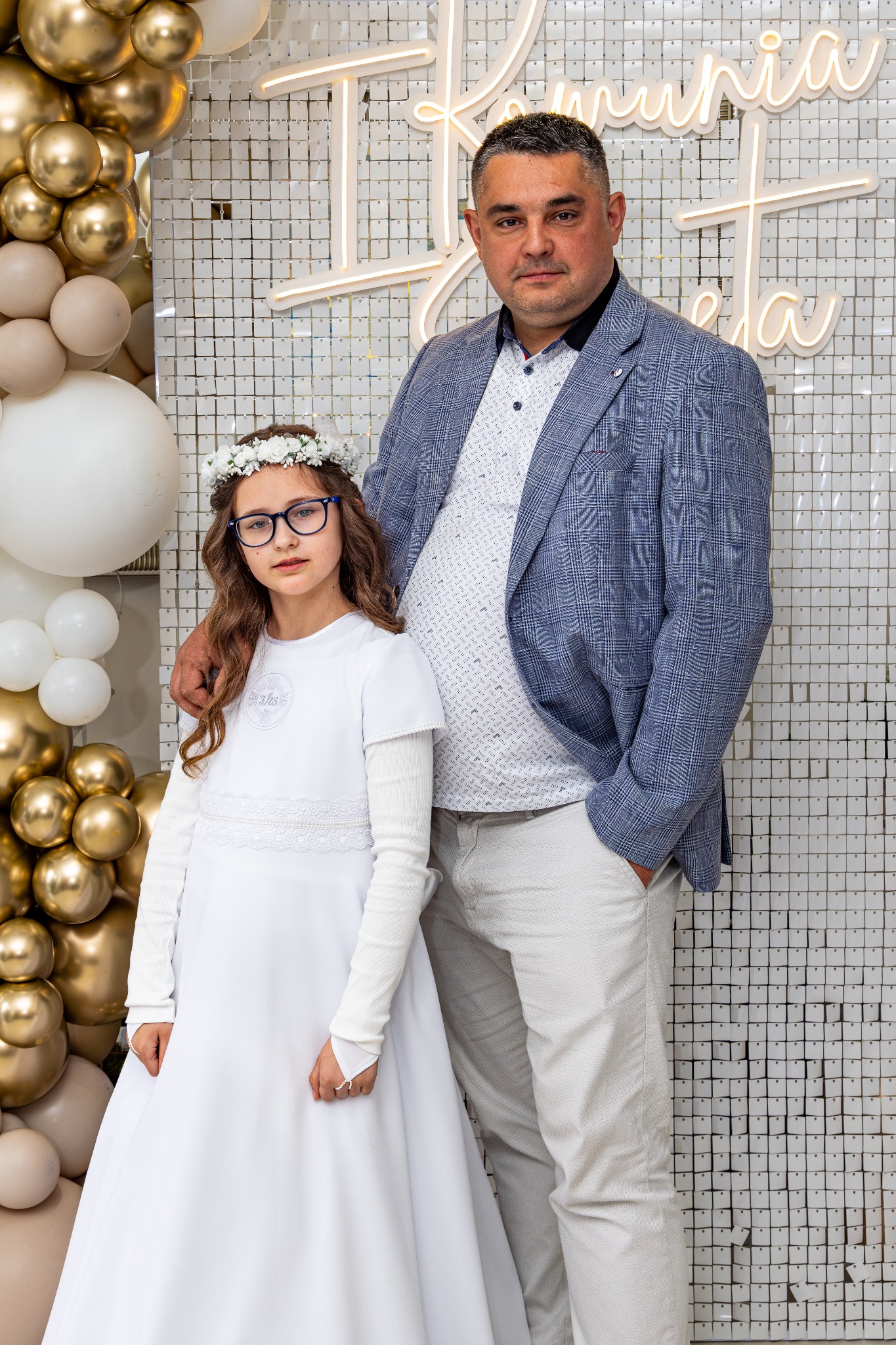 Portret dziewczynki w białej sukience komunijnej z tatą na tle ściany z dekoracją balonową i neonowym napisem 'Komunia Święta'.
