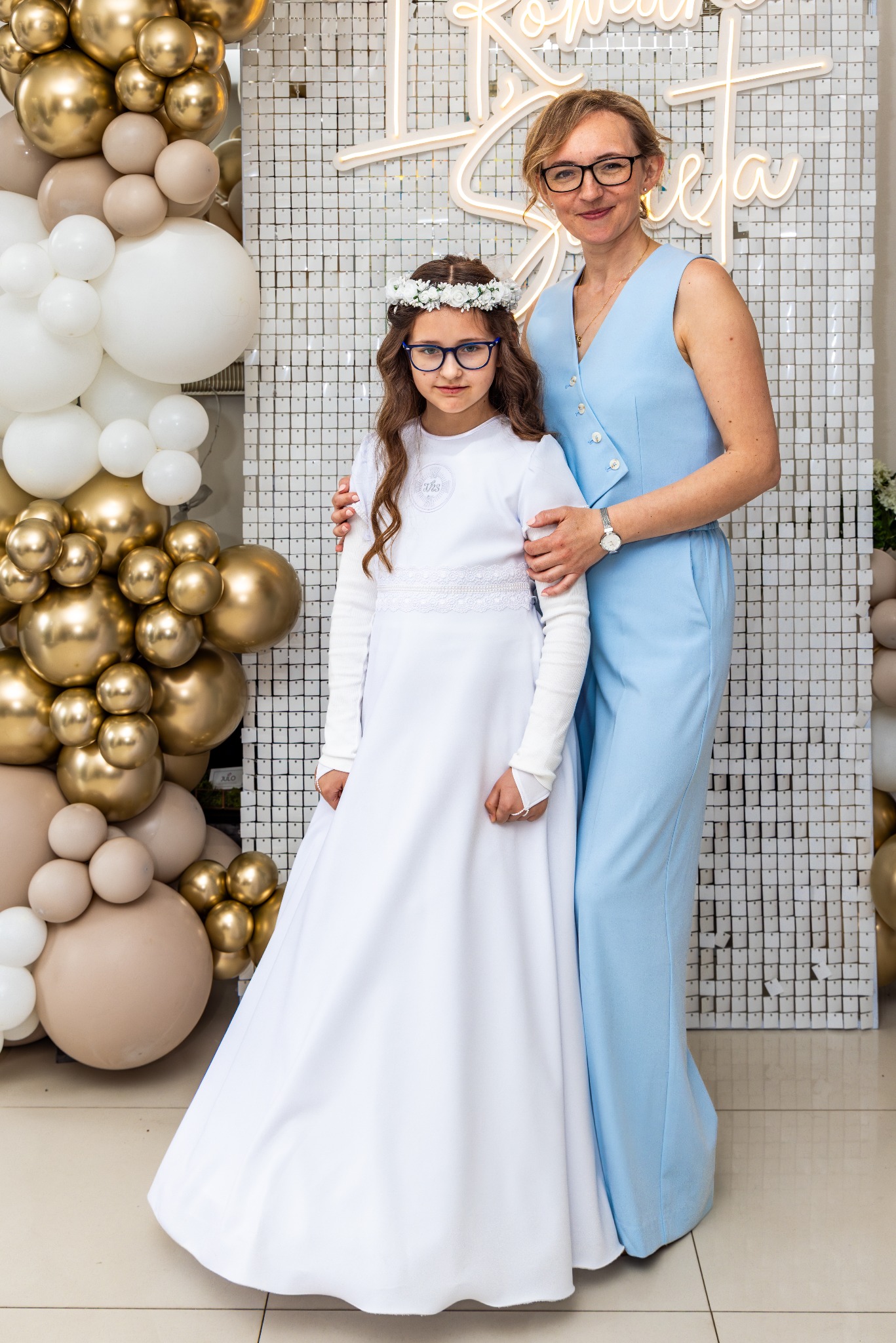 Dziewczynka w białej sukience komunijnej z mamą na tle dekoracji z balonów i błyszczącej ścianki, ujęcie w pełnej sylwetce, delikatne oświetlenie.